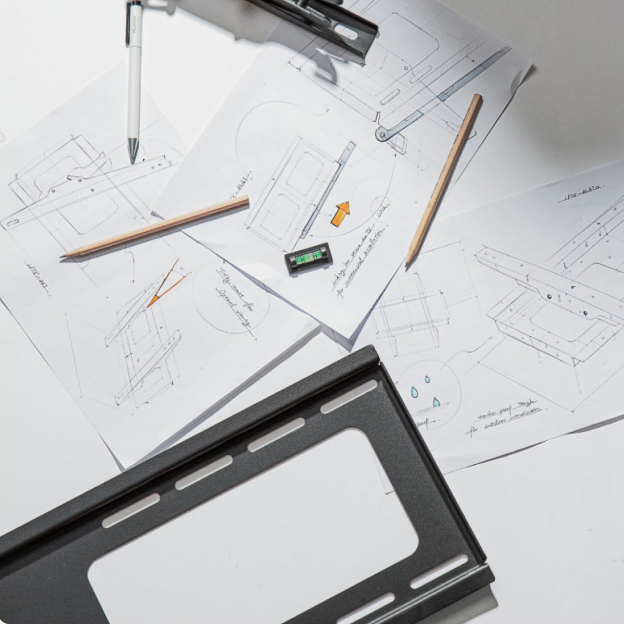Technical sketches of monTEK mounting hardware with pencils, pen, level, and metal bracket on a white workspace